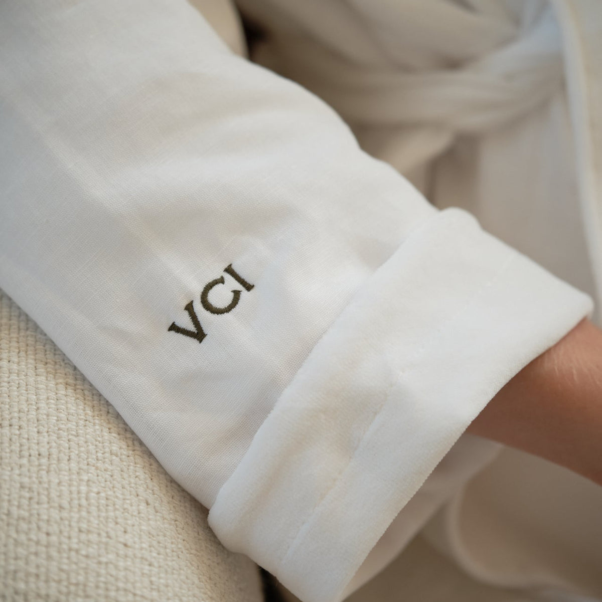 A close-up of a white fabric garment, likely a robe or wrap, with the cuff turned up to reveal the embroidered text 'VCI' in dark brown or black thread. A small portion of a person's arm is visible at the cuff.