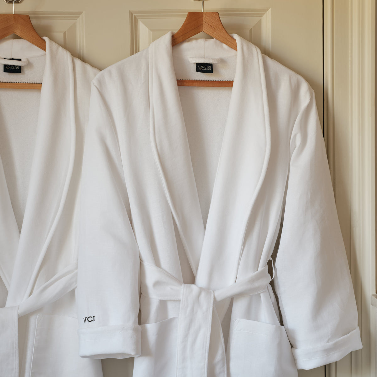 A white bathrobe hanging on a wooden hanger against a cream-colored paneled door, with a second robe partially visible behind it.