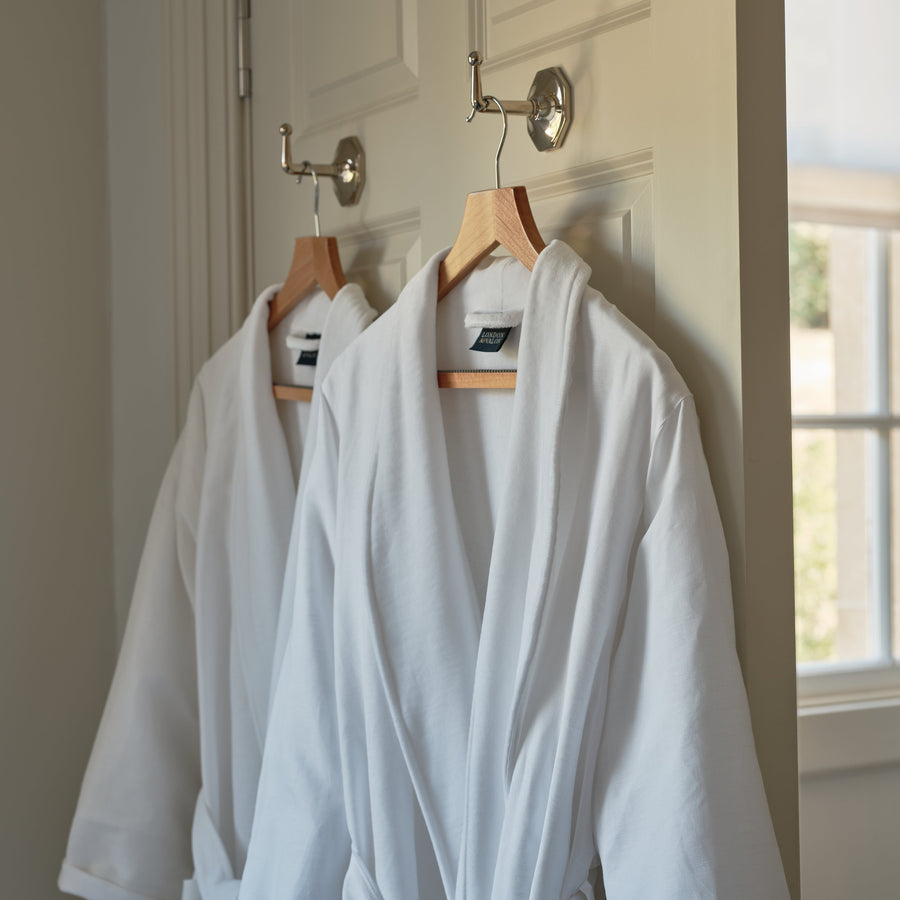 Two white bathrobes hanging on wooden hangers from a silver door hook on a white paneled door, with a window visible in the background.