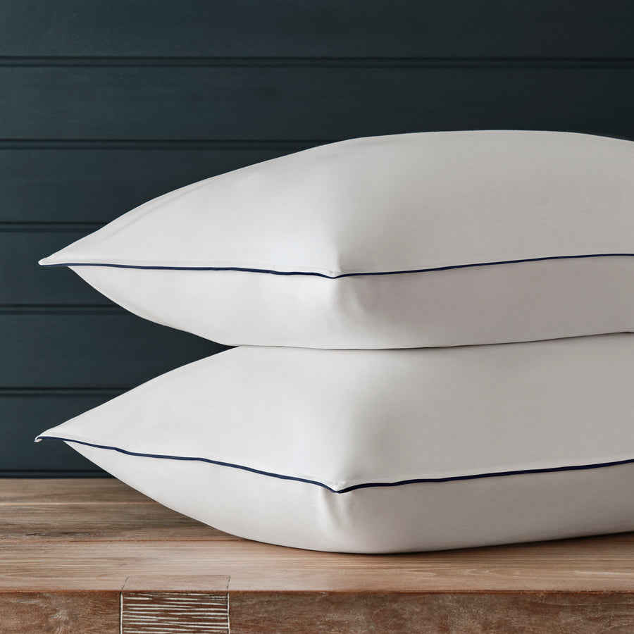 Two white pillows stacked on a wooden surface against a dark paneled wall, each trimmed with a thin dark blue piping detail.